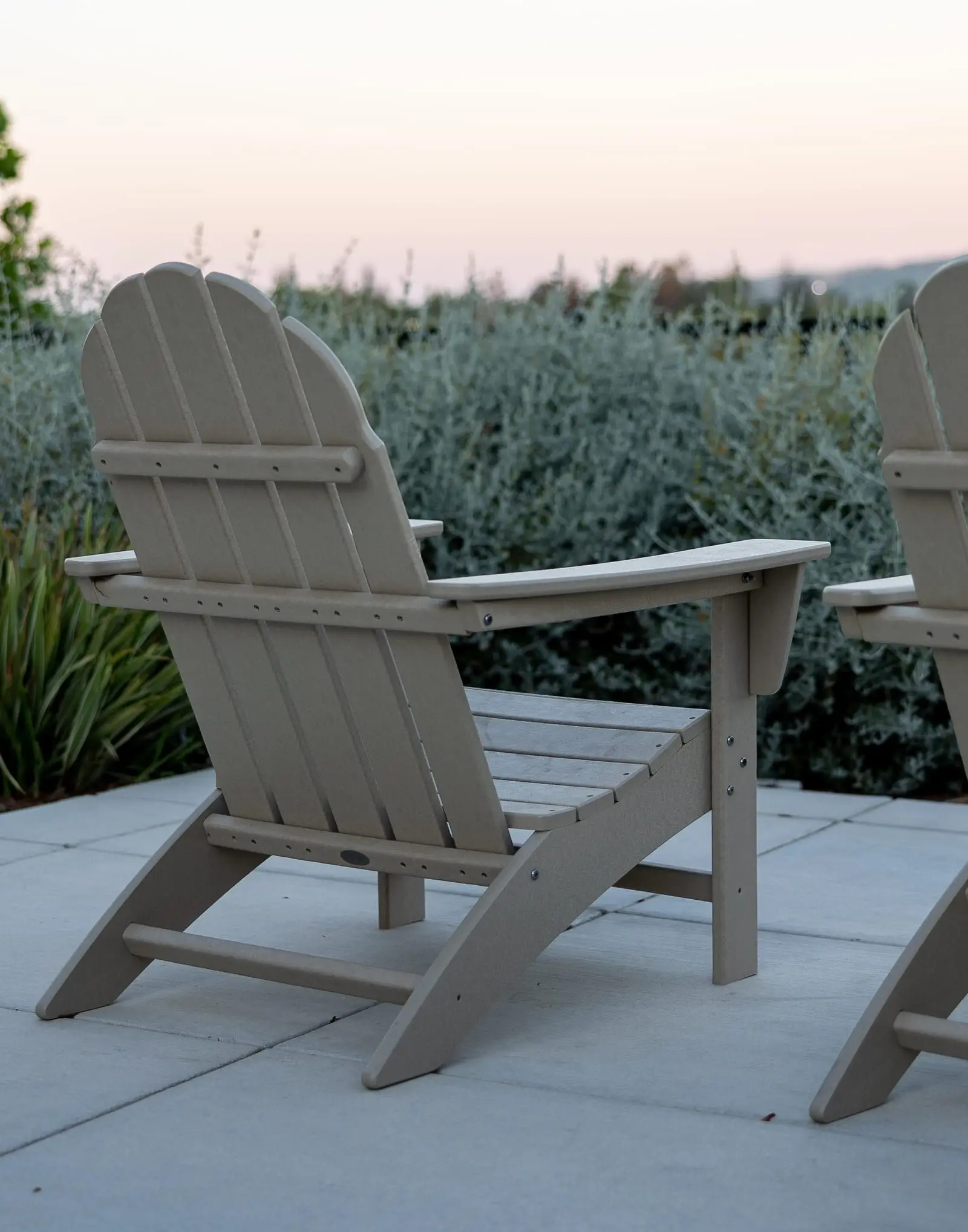 A timber garden chair on large pale pavers at dusk, facing a bed of grey-green shrubs and strappy plants