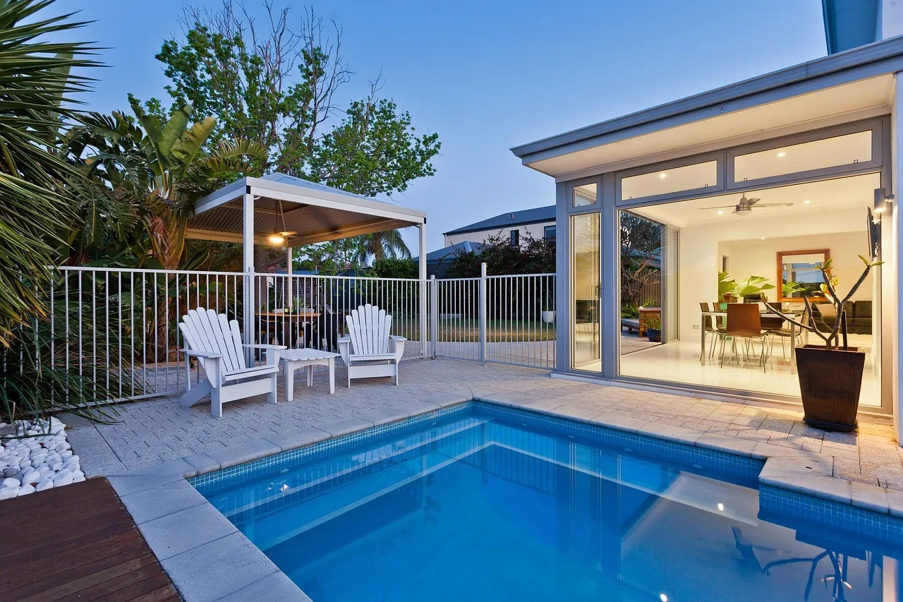 A paved pool surround at dusk with two white timber chairs, a steel-roofed pergola behind a white pool fence and a lit glass-walled room