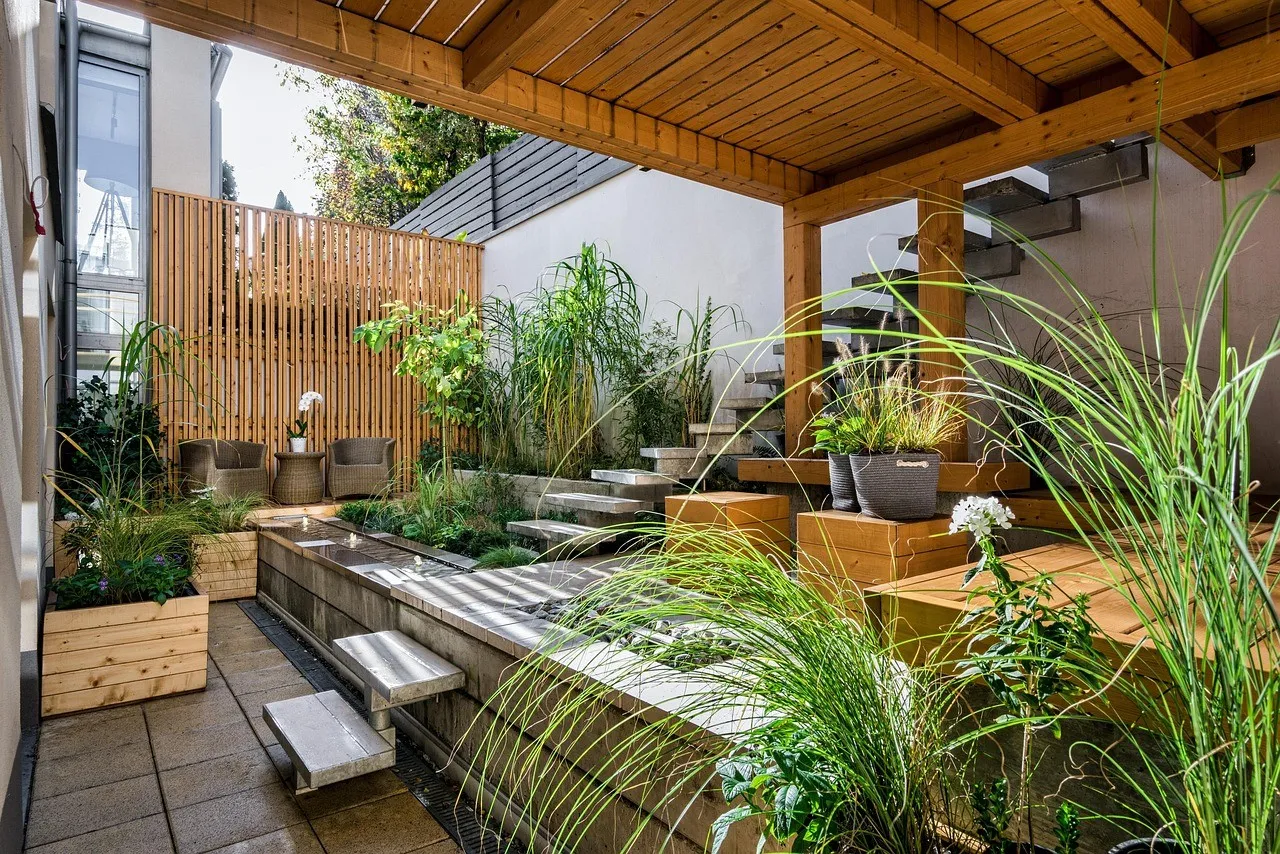 A paved courtyard under a timber pergola, with a raised water feature, a timber slat screen and planter boxes of grasses