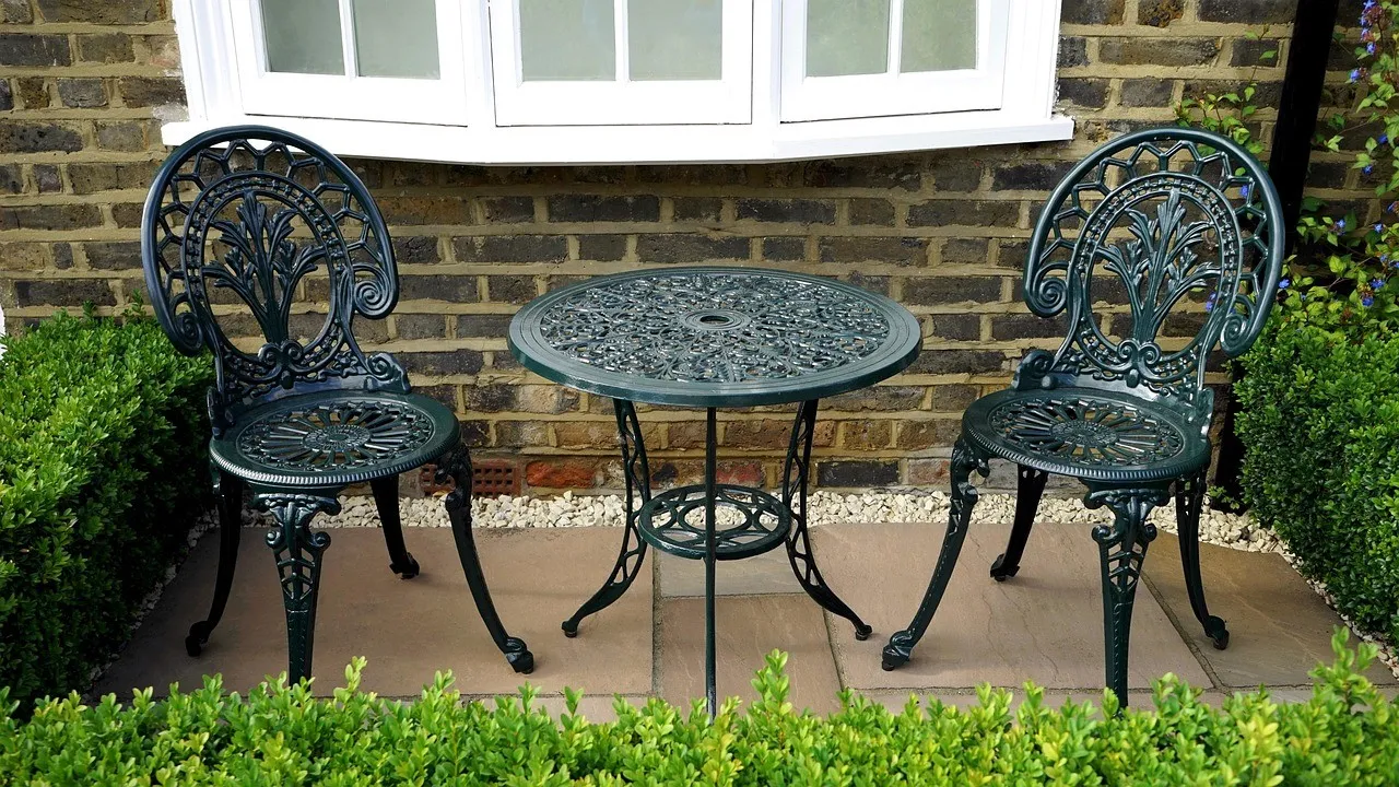 A small sandstone-paved terrace edged with clipped box hedging, with a dark green cast-iron table and two chairs against a brick wall